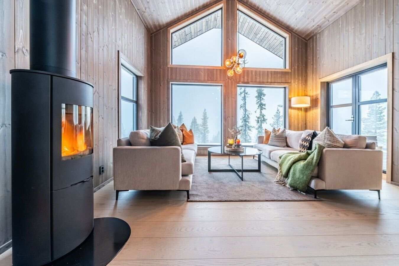 A modern living room with a wood-burning stove, two sofas, a coffee table, and large windows offering a view of trees.