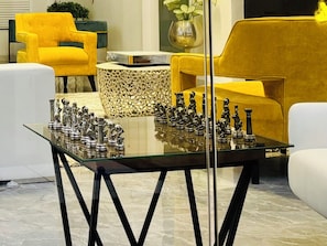 Una sala de estar moderna con una mesa de centro de vidrio que muestra un juego de ajedrez, rodeada de sillones tapizados en amarillo y un reposapiés estampado.