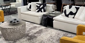 Uma sala de estar moderna com um sofá branco, almofadas decorativas, uma mesa de centro de vidro e um tapete estampado.