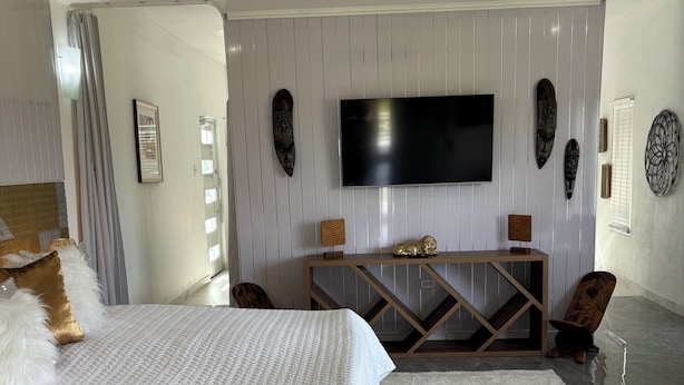A modern bedroom with a large bed, a TV mounted on the wall, and a ceiling fan. - Villa Near Negril | 5BR Private Pool & Spa | Caribbean Family & Group Retreat (LITTLE LONDON)