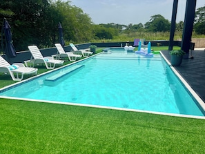 Una piscina rectangular con borde de azulejos, rodeada de sillones blancos con cojines azules, y un área de césped con árboles al fondo.