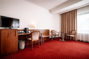 A hotel room with a TV, a desk with a lamp, two chairs, a small table, and a red carpet.