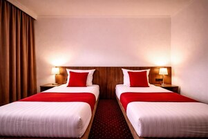 A hotel room with two beds, each with a red pillow and white bedding. There are two bedside lamps and a wooden headboard.