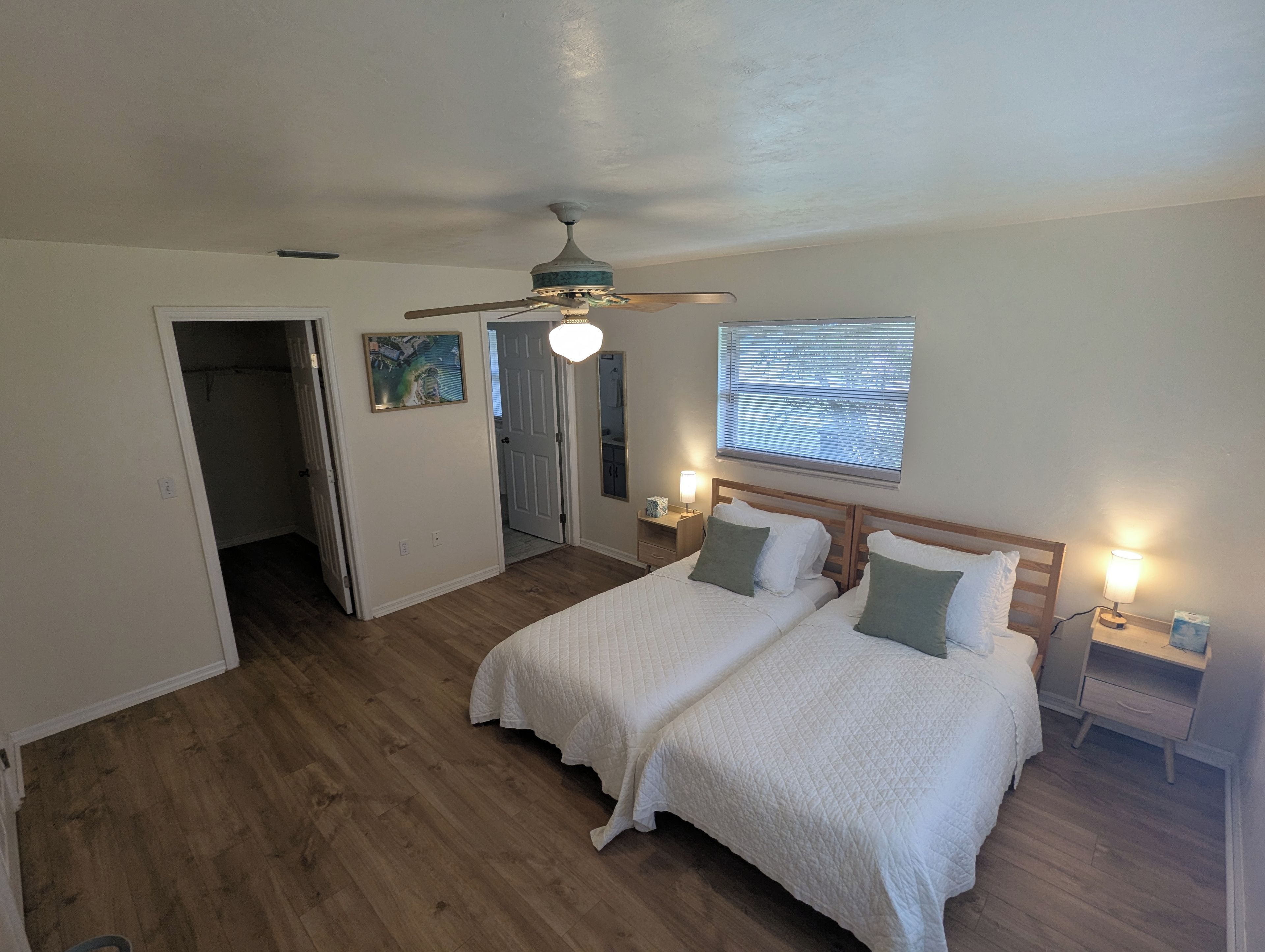 A bedroom with two beds, a ceiling fan, a window with blinds, and a door leading to another room.