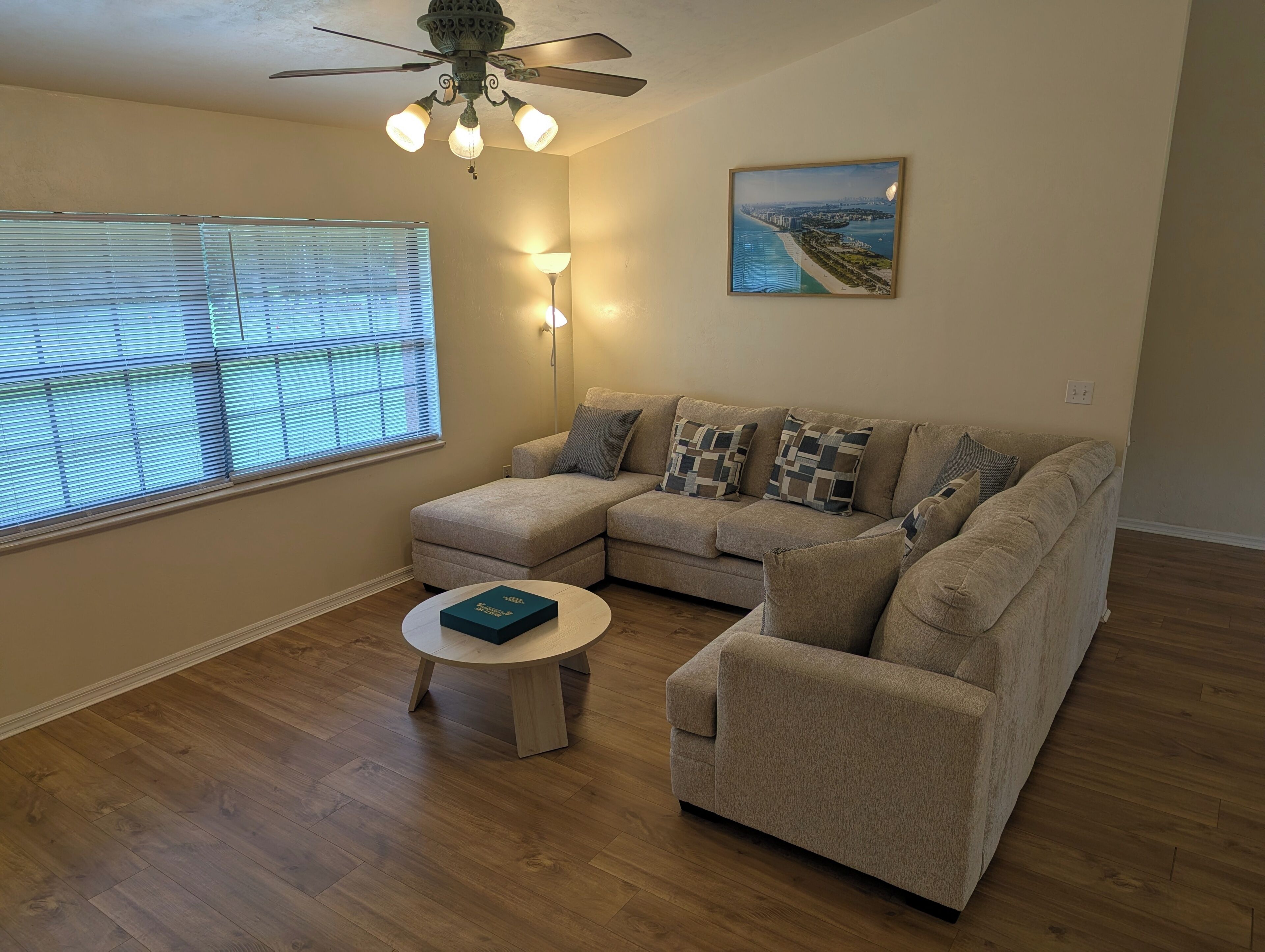 A living room with a sectional sofa, a round coffee table, a ceiling fan, a floor lamp, and a framed picture on the wall.