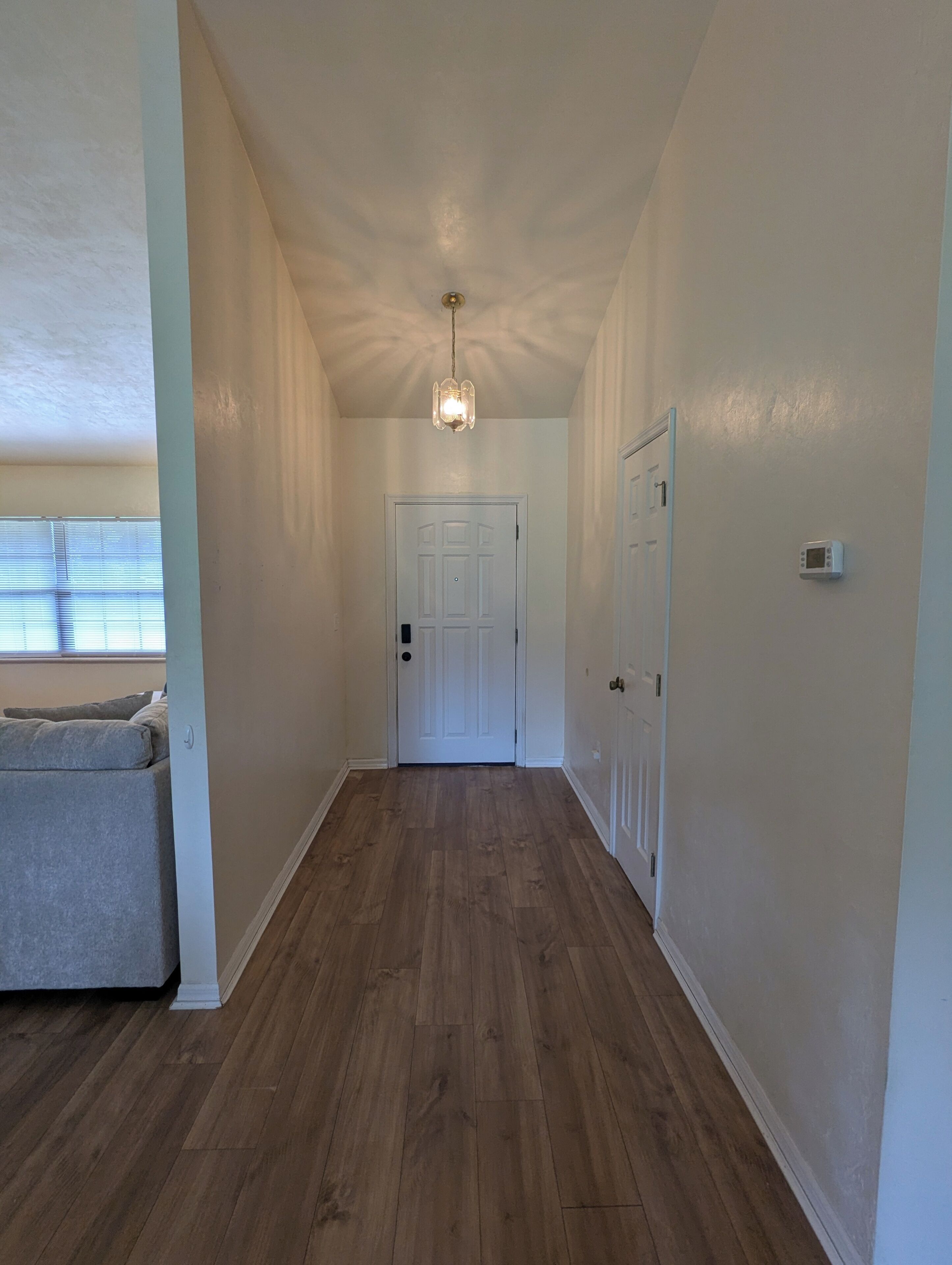 A hallway with wooden flooring, white walls, a white door at the end, and a ceiling light fixture.