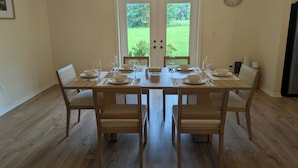 A dining area with a table set for four, chairs, a clock on the wall, and a view of a green lawn through a glass door.