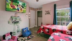 Ein Schlafzimmer mit Fernseher, Deckenventilator, einem fenster mit Vorhängen, einer Tür und einer Bettdecke im Mickey Mouse Stil.