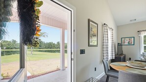 A room with a view of a grassy area, a dining table with chairs, and a window with a curtain. - Come stay at peaceful Timberland Cottage, a fully furnished 3 bedroom property. (Claxton)