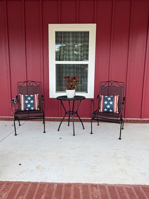 Two metal chairs with American flag cushions, a small round table, and a potted plant with red flowers, all set against a red wall with a white-framed window.
