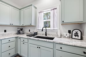 Una cocina con armarios blancos, un lavamanos, una cafetera y una ventana con cortinas.
