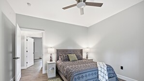 A bedroom with a bed, bedside lamps, a nightstand, a floor lamp, a ceiling fan, and a door leading to another room.