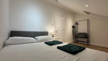 Ein Schlafzimmer mit Bett, Kissen, einem Nachttisch mit Lampe, einer Tür und einem schwarzen Metallregal mit Matratze.
