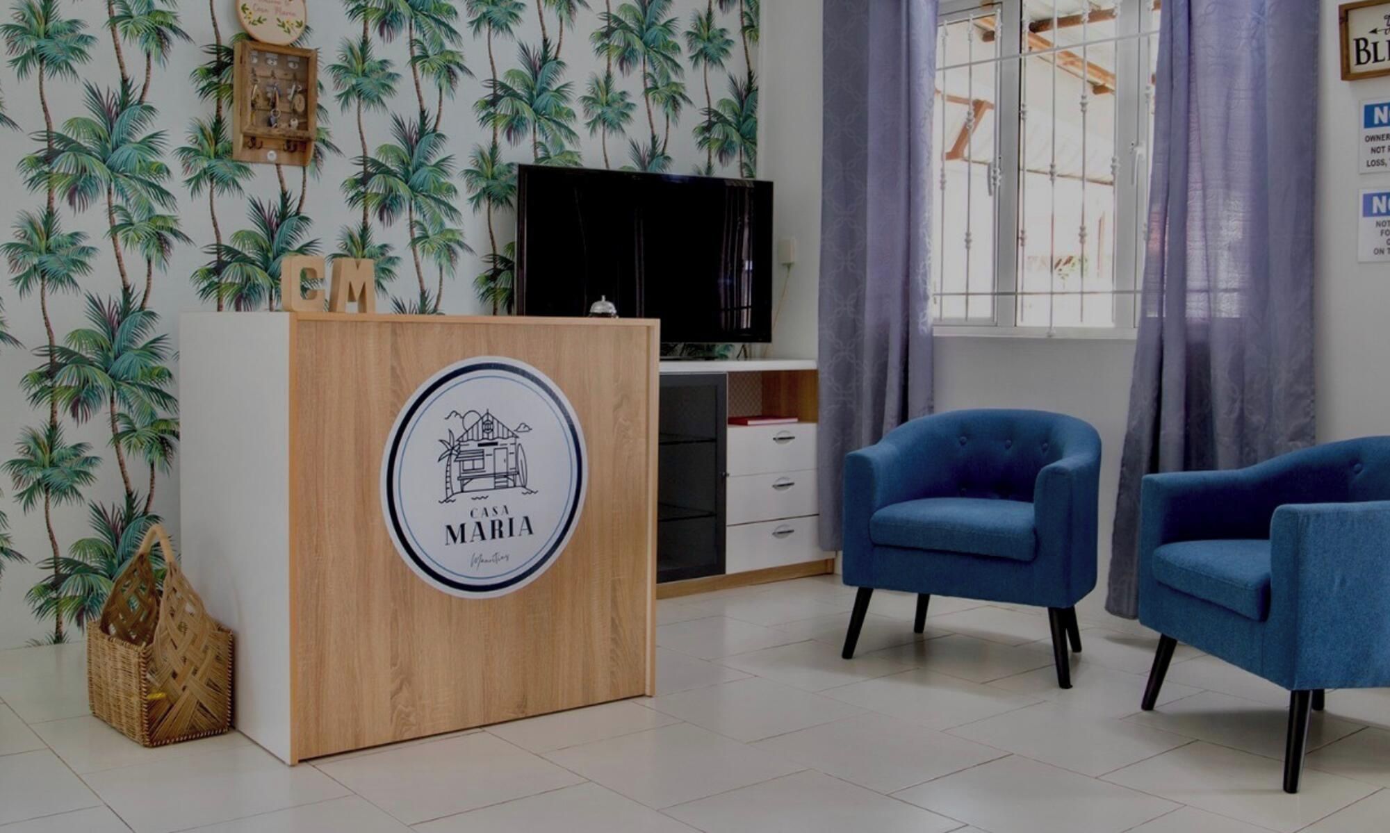 A room with a wooden reception desk, two blue armchairs, a television, and a window with curtains.