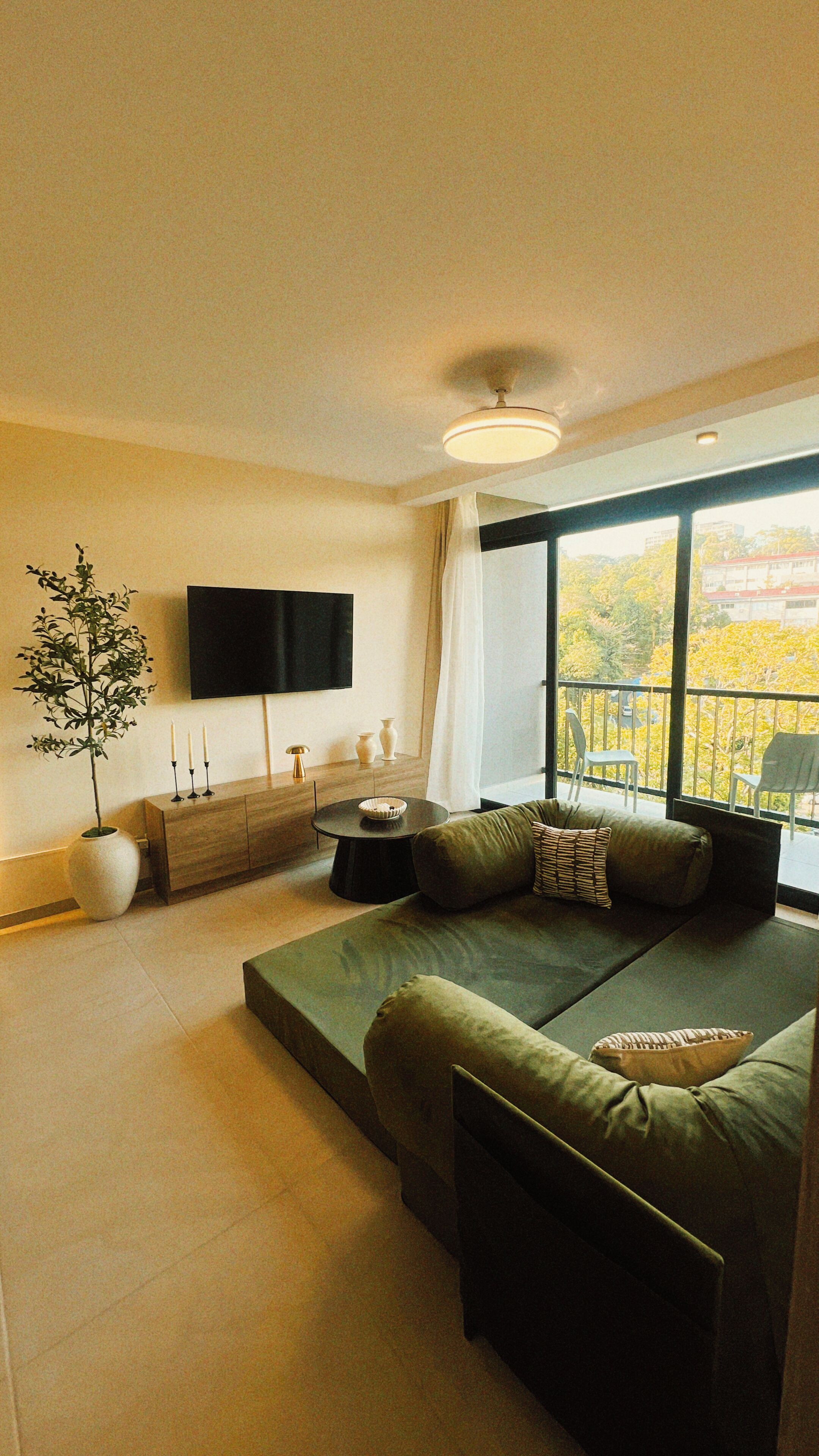 A modern living room with a green sofa, a flat-screen TV, a potted plant, and a view of a yellow autumn landscape.
