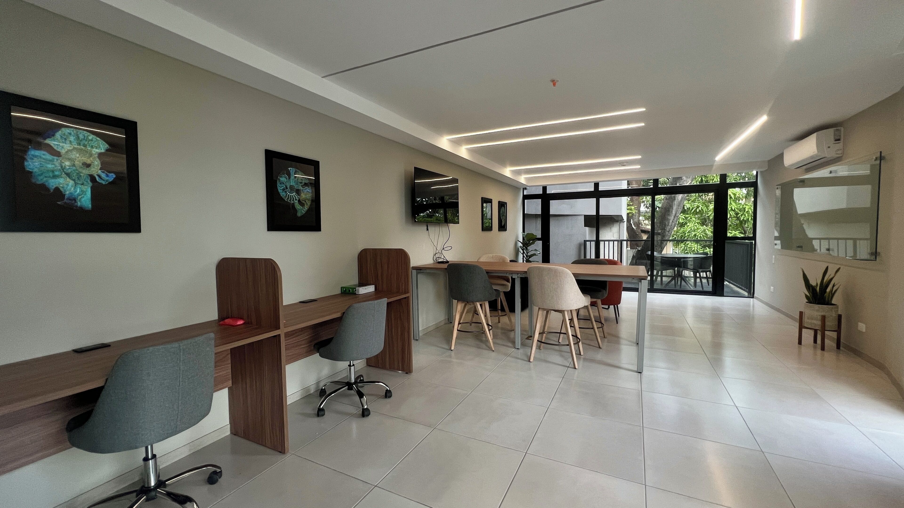 A modern office space with long wooden desks, ergonomic chairs, and a large window providing a view of the outdoors.