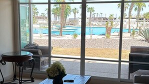 A room with a large window, a ceiling fan, a wooden coffee table with a vase, and a view of a pool and palm trees outside.
