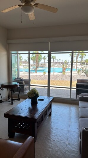 Um quarto com uma grande janela, um ventilador de teto, uma mesa de centro de madeira com um vaso, e vista para uma piscina e palmeiras lá fora.
