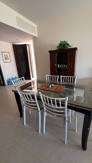Uma sala de jantar com mesa de vidro, cadeiras brancas, um armário de madeira escura e uma porta que dá para outro cômodo.