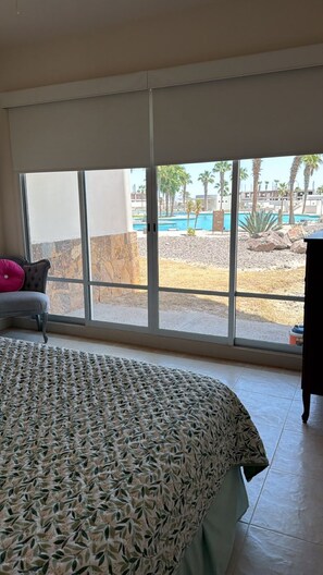 Quarto com uma janela grande, uma cama com colcha estampada, uma cadeira e vista para a área da piscina.