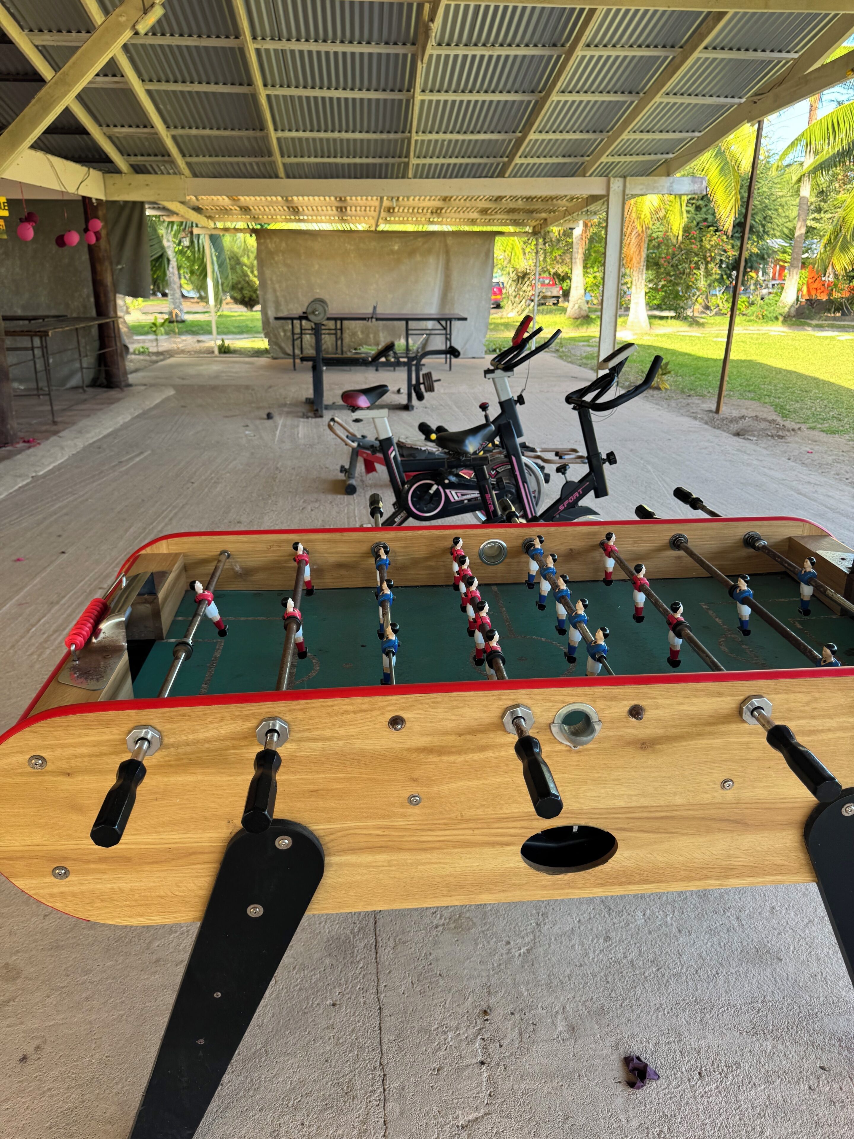 A foosball table with a metal frame and a wooden playing surface, located in an outdoor covered area with a concrete floor.
