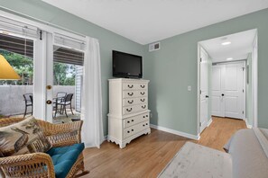 A room with a white dresser, a television on top, wicker furniture, and a view through a glass door. - Island Ease Villa Golf, Beach, Marina Nearby (Hilton Head Island)
