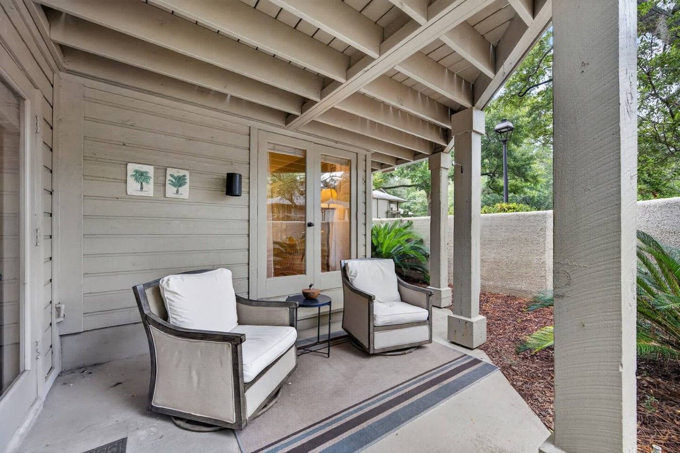 A covered porch with two chairs and a small table, a door with glass panels, and a wall with framed pictures.