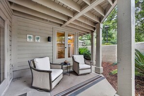 A covered porch with two chairs and a small table, a door with glass panels, and a wall with framed pictures. - Island Ease Villa Golf, Beach, Marina Nearby (Hilton Head Island)