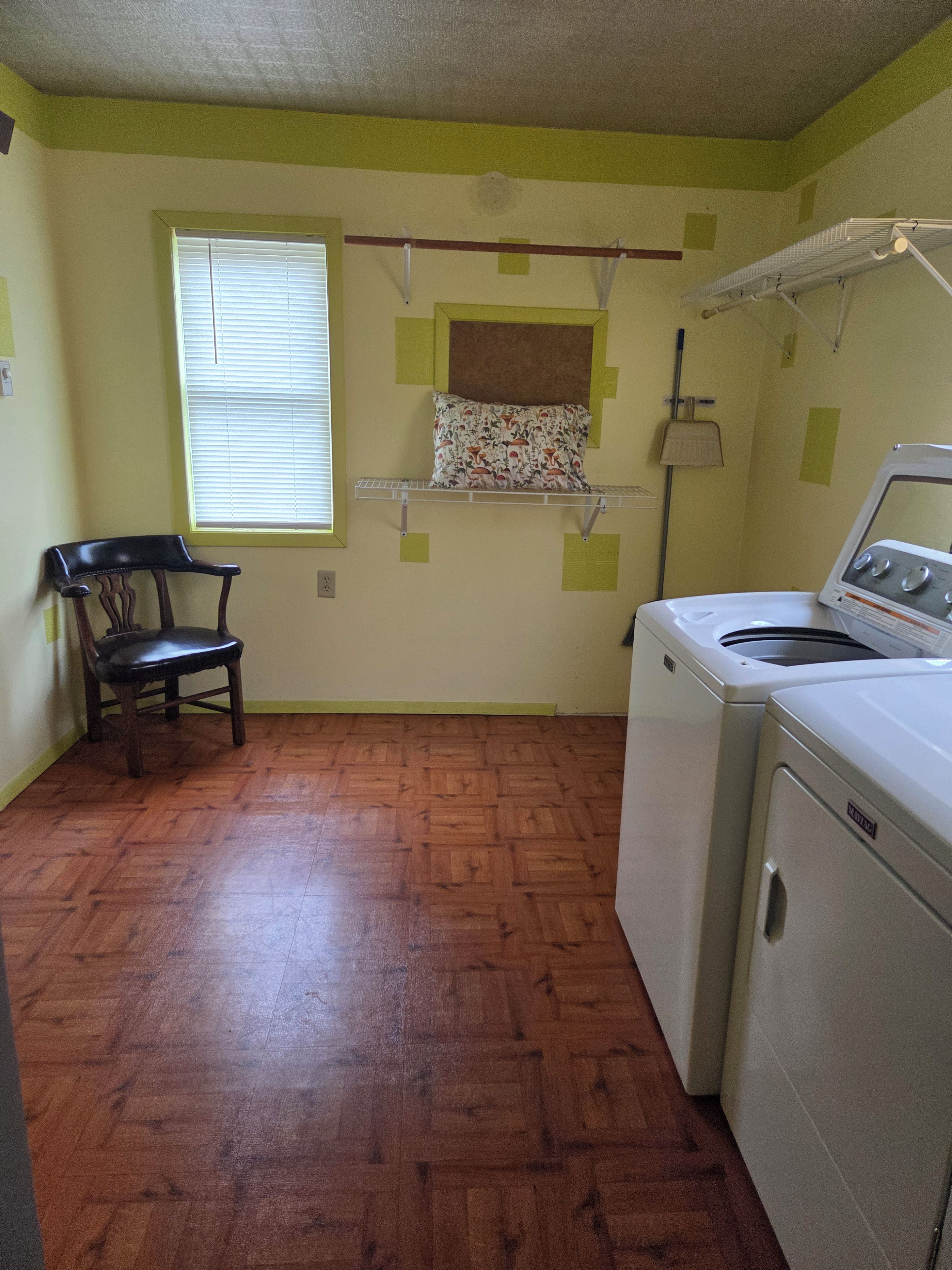 A laundry room with a washing machine, a chair, and a window with blinds.