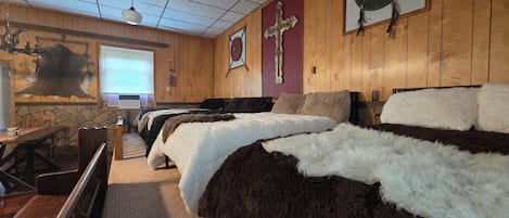 A room with wooden walls, a cross, a shield, a stone wall, a bed with a fur blanket, a wooden bench, and a stone fireplace.
