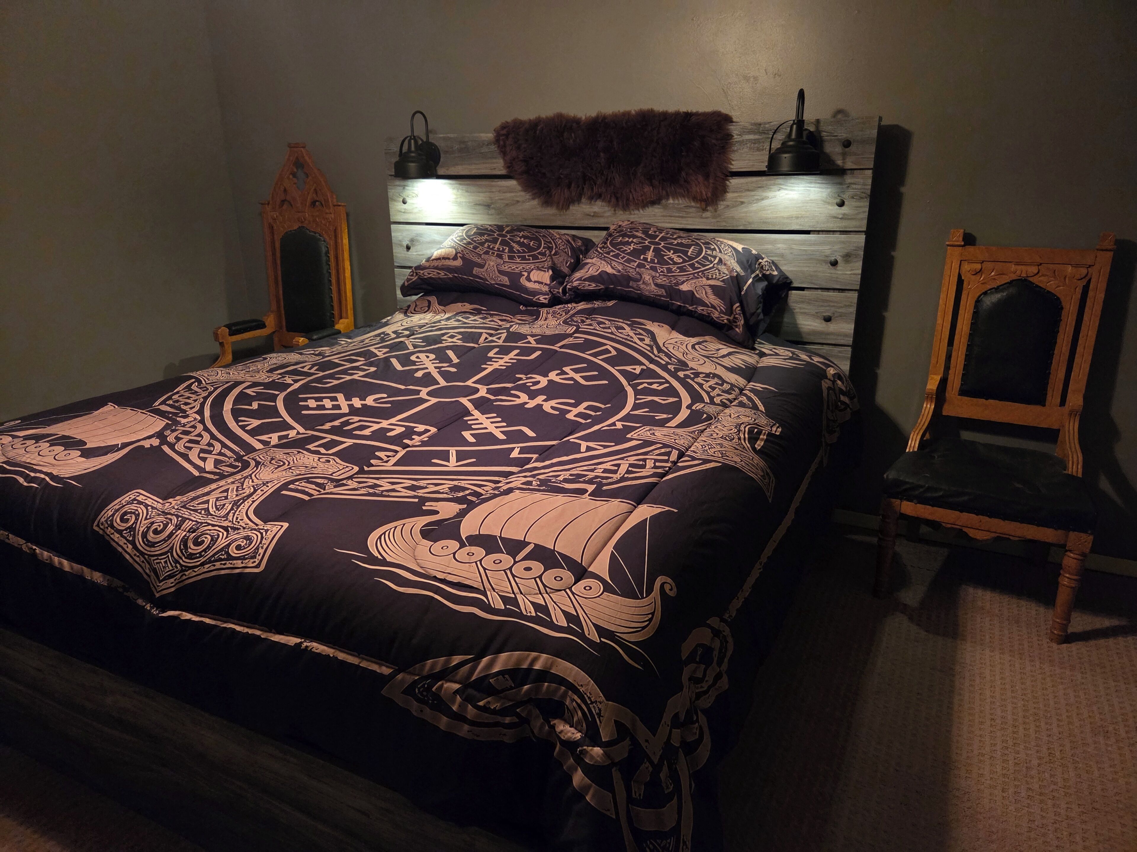 A bed with a patterned bedspread, a wooden headboard, a chair, and a wall-mounted lamp.