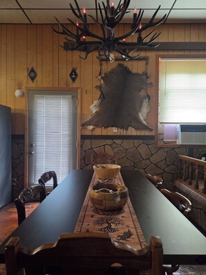 A dining area with a long table, wooden chairs, a chandelier with antlers, a fur rug, and a stone wall. - Beautiful Viking "Long Hall" lodge with AC in charming Waubay SD. (Waubay)