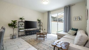 A modern living room with a flat-screen TV, a sofa, a coffee table, and a large window with a view of a building. - 4BR Storey Lake Resort Near Disney Private Pool (Kissimmee)