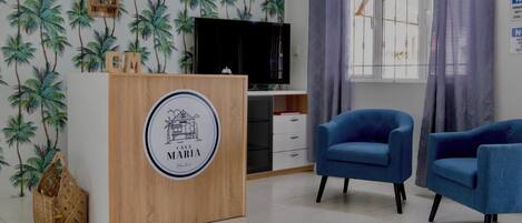 A room with a wooden reception desk, two blue armchairs, a television, and a window with curtains.