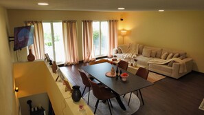 Un salon comprenant un canapé, une table à manger avec des chaises, un téléviseur fixé au mur et une fenêtre avec des rideaux.