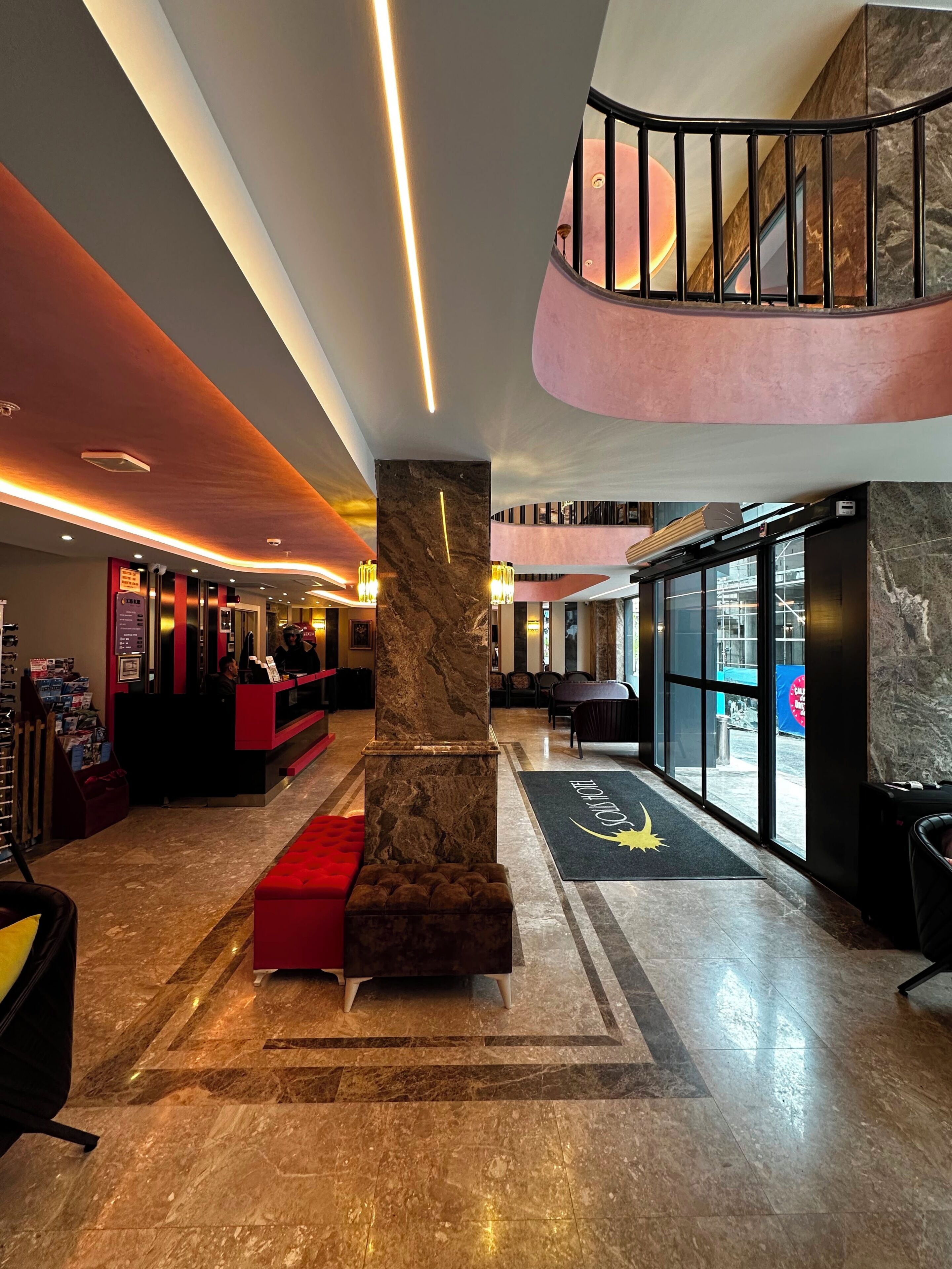 A hotel lobby with a reception desk, a red bench, a marble pillar, and a staircase leading to an upper level.