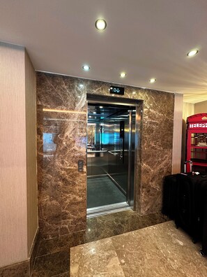 Un ascensor con pantalla digital, paredes de mármol y una silla roja al fondo.