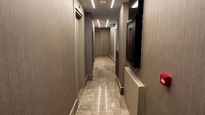 Un pasillo de hotel con piso de mármol, iluminación empotrada, un televisor de pantalla plana en la pared y una caja roja de alarma contra incendios.