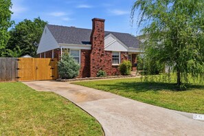 A residential house with a brick chimney, a wooden fence, and a well-maintained lawn. - Clementine Cottage - By Fairgrounds, Kid-Friendly - Stay Smiley Rentals (Oklahoma City)