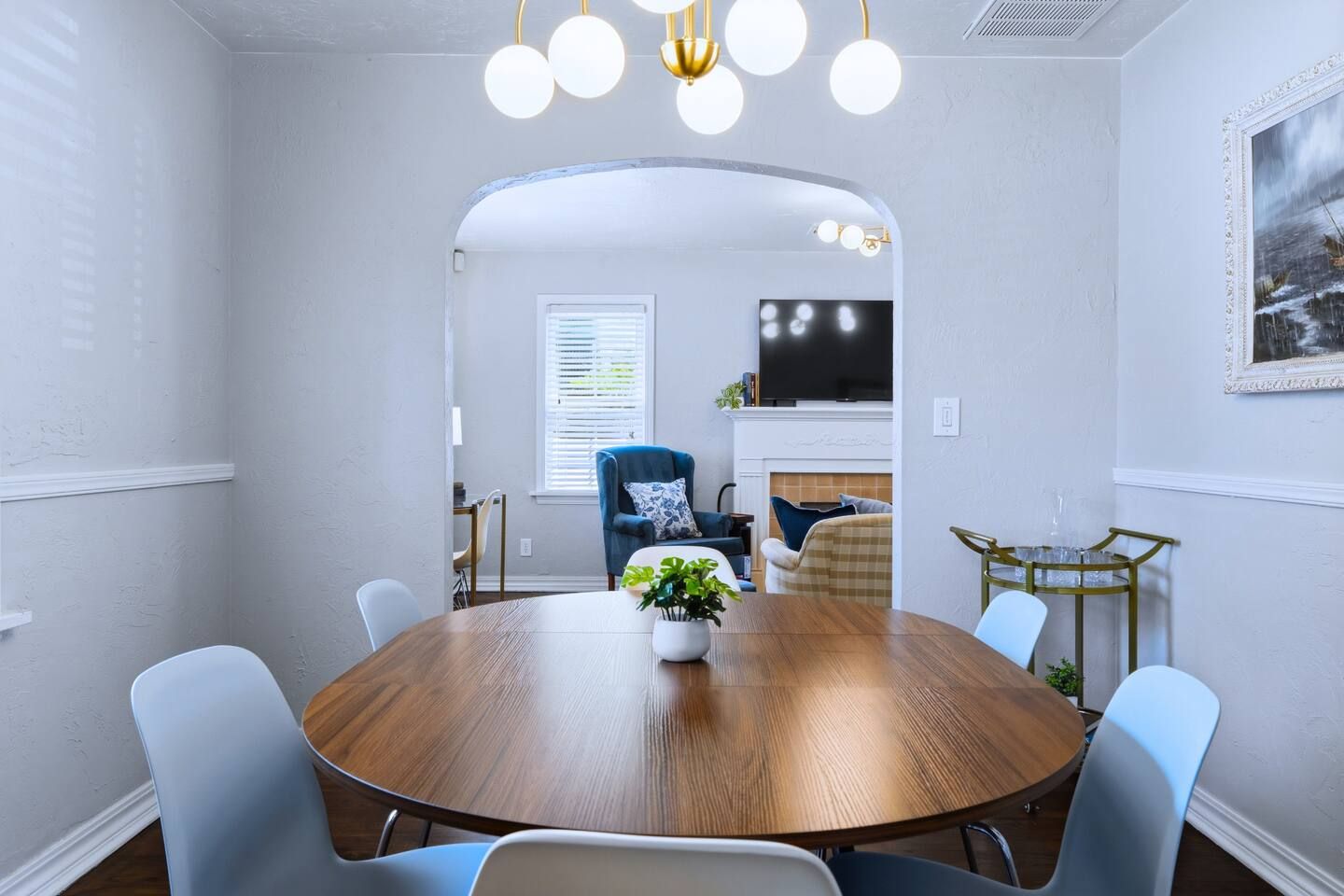 Un comedor con una mesa redonda de madera, seis sillas, una planta en maceta y un televisor al fondo.