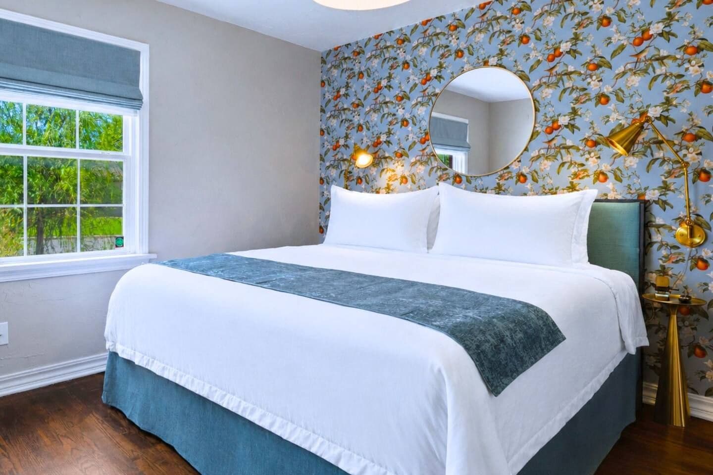 A neatly made bed with white linens and a blue accent, a round mirror above it, and a floral wallpaper pattern on the wall.