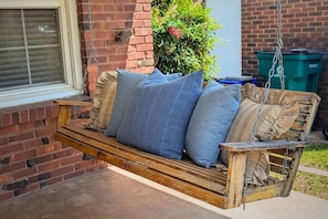 A wooden swing with cushions, a brick wall, and a green trash bin in the background. - Clementine Cottage - By Fairgrounds, Kid-Friendly - Stay Smiley Rentals (Oklahoma City)