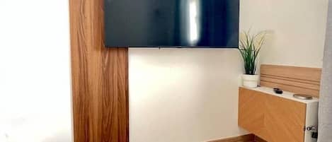 Una habitación con una televisión de pantalla plana montada en la pared, un armario de madera, una planta pequeña en maceta y un reloj en la pared.