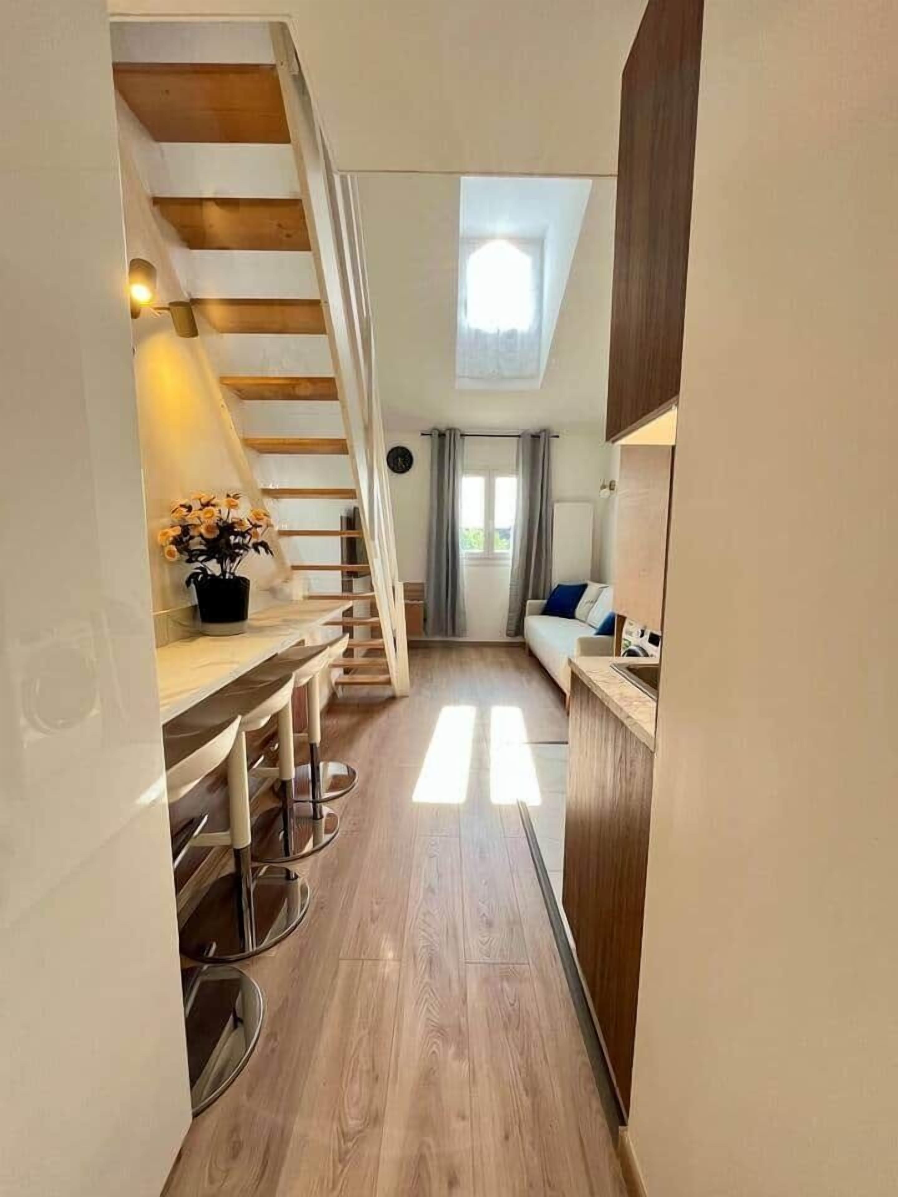 Een smalle gang met een houten trap naar een slaapkamer op een entresol, een klein keukenhoekje en een zithoek met uitzicht op de omgeving.
