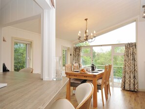 A dining area with a wooden table, chairs, and a chandelier. There are windows with curtains, and a view of greenery outside.