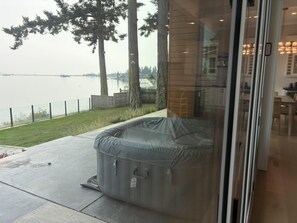 Une pièce entièrement vitrée donnant sur un lac, avec un jacuzzi et un patio entouré d’une clôture et bordé d’arbres.