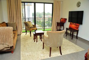 Ein Wohnzimmer mit einer Couch, Sesseln, einem Couchtisch, einem Fernseher und Blick auf einen Park und Gebäude.