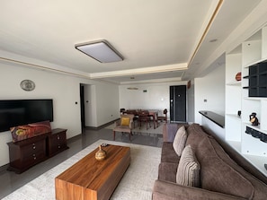 En moderne stue med et fladskærms-tv, et træsofabord, en brun sofa og et spiseområde med bord og stole.