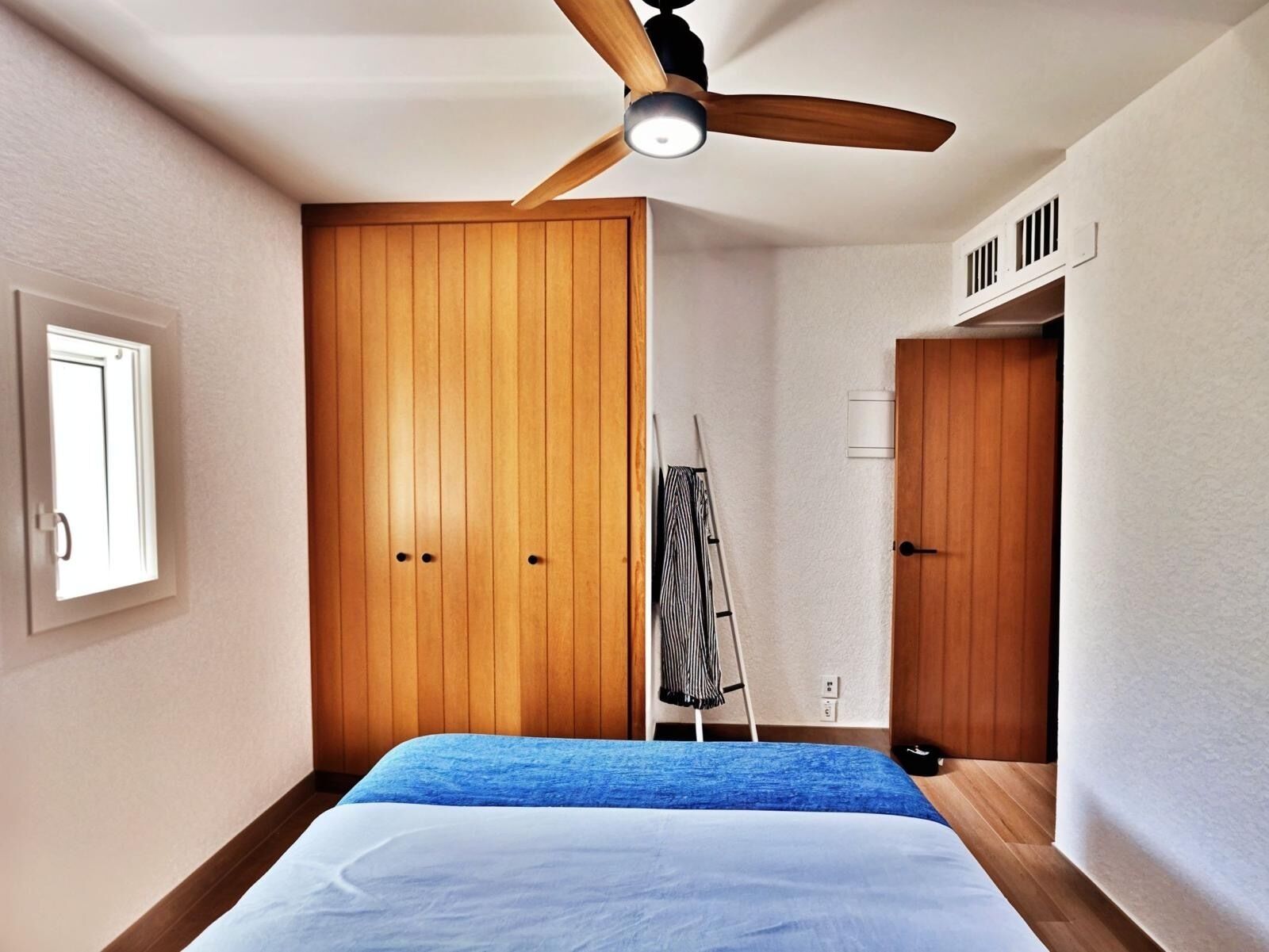 A bedroom with a bed, wooden wardrobe, ceiling fan, and a window.