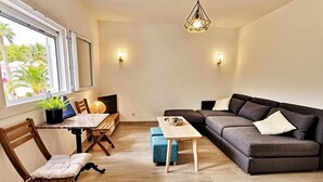 Olohuone, jossa on sohva, ruokapöytä tuoleineen, televisio ja ikkuna, josta avautuu näkymä palmuille.
